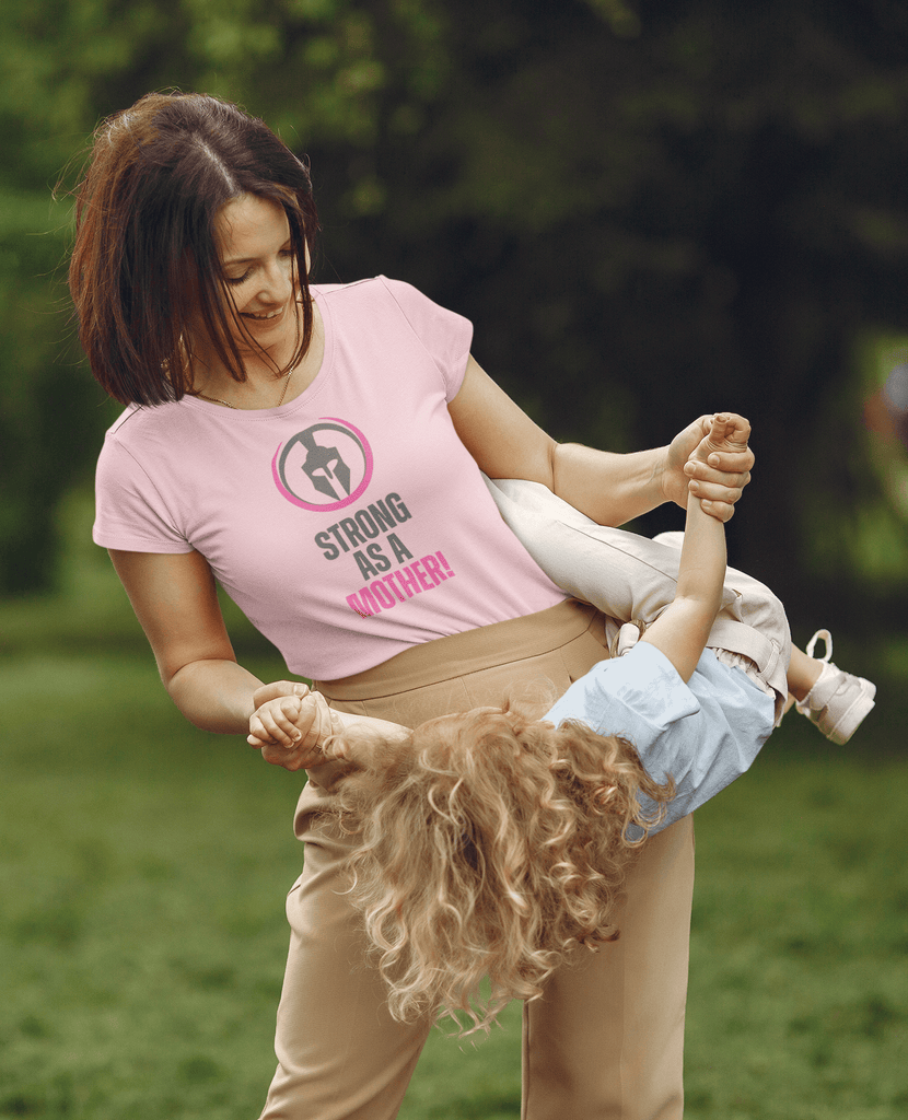 Woman wearing 'Strong as a Mother' pink tee from Female Warrior Collection, playing with child outdoors.